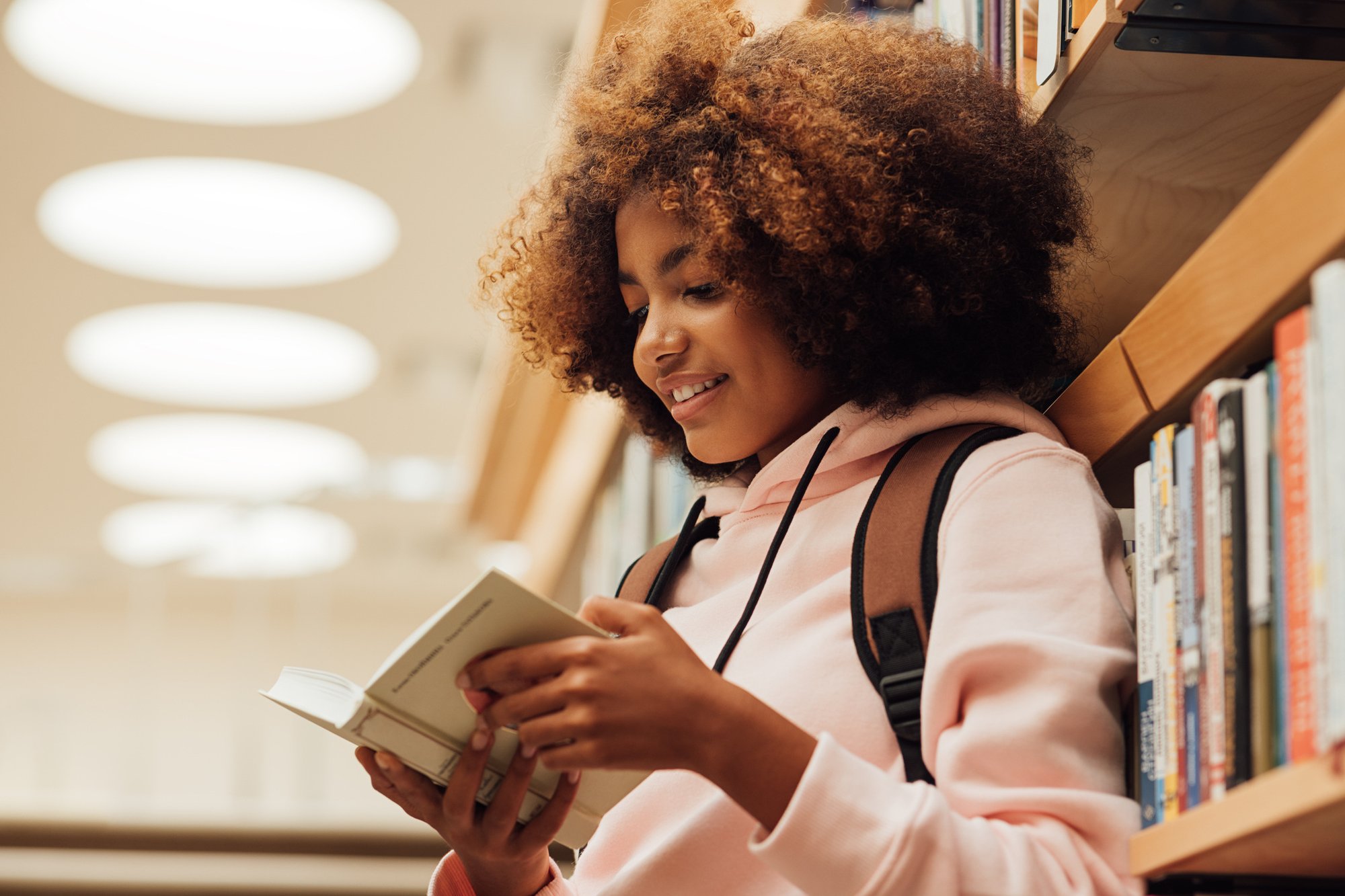 Smiling middle school child reading a book while leaning against a bookshelf in a library