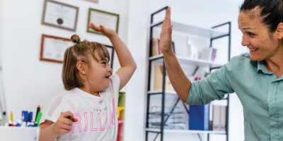 Classroom setting in which woman in green button down shirt high fives child with down syndrome in a white shirt Classroom setting in which woman in green button down shirt high fives child with down syndrome in a white shirt