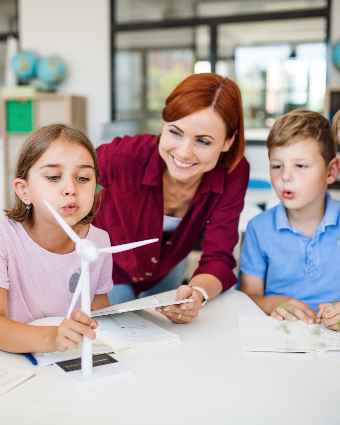 smiling teacher with two students who are blowing on a mini wind turbine in a classroom